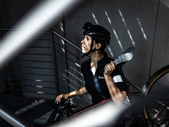 A white woman wearing a black shirt and helmet carries a bicycle with a 3d printed saddle as she climbs stairs in a stairwell with some sunlight streaming through.