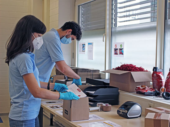 A young woman and a young man wearing protective masks and light blue polo shirts are packing small boxes while standing at a desk.