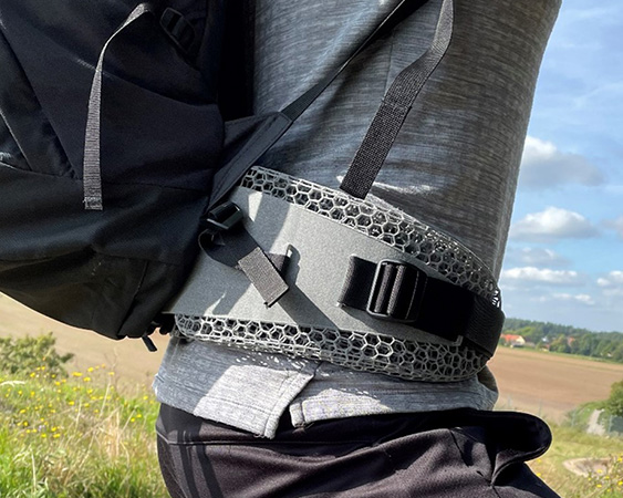 The waist of a man wearing a gray t-shirt with the lattice structure padding of the backpack tied around it. In the background a field, trees and blue sky with white clouds.