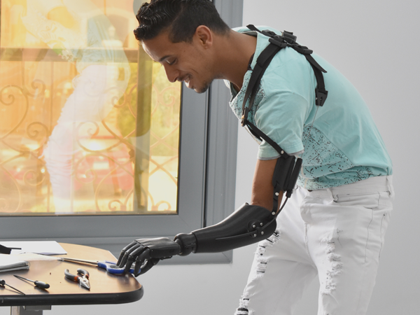 A young man of Color with short hair and a turquoise t-shirt wears a black prosthetic arm and reaches for a pair of scissors lying on a table.